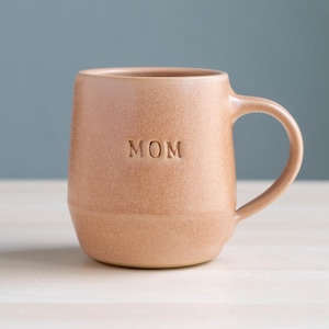 Peut inclure: Une tasse en céramique beige avec le mot "MOM" en relief sur le devant. La tasse a une forme arrondie avec une anse sur le côté. La tasse est posée sur une surface en bois clair, sur fond bleu-gris.