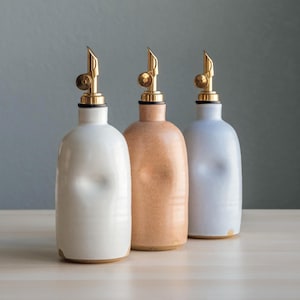 Op de afbeelding: Drie keramische olieflessen in crème, perzik en lichtblauw, elk met een goudkleurige tuit en een zwarte ring rond de hals. De flessen staan op een lichtgekleurd houten oppervlak tegen een gedempte grijze achtergrond.