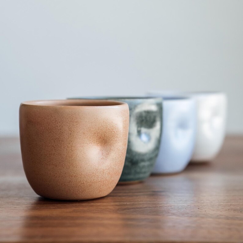 Pu&ograve; includere: Quattro tazze in ceramica di diversi colori e texture. Le tazze sono allineate su una superficie di legno. La tazza a sinistra &egrave; di colore marrone chiaro con una superficie testurizzata. La tazza successiva &egrave; di colore grigio scuro con un design a spirale bianco. La terza tazza &egrave; di colore blu chiaro con una superficie liscia. L'ultima tazza &egrave; di colore bianco con una superficie testurizzata.
