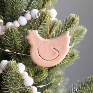 May include: A light brown chicken-shaped Christmas ornament hanging on a green Christmas tree. The ornament has a simple, smiling face and is suspended by a gold string. White pom-pom garland and string lights decorate the tree.