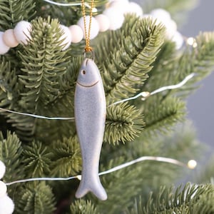 May include: A ceramic fish ornament in shades of gray hangs from a gold string on a Christmas tree. The fish has a simple design with a small eye and a curved mouth. The tree is decorated with white pom-pom garland and string lights.