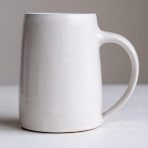 Op de afbeelding: Een witte keramische mok met een afgerond lichaam en een groot handvat. De mok is gemaakt van een gladde, matte afwerking.