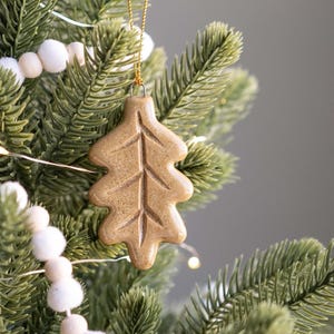 May include: A tan, leaf-shaped Christmas ornament hangs from a gold chain on a green Christmas tree. The ornament has detailed veining. White pom-pom and wooden bead garland and fairy lights decorate the tree.