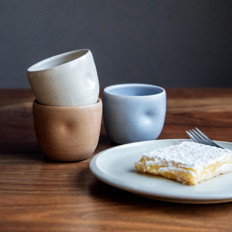 Pu&ograve; includere: Tre tazze in ceramica di diversi colori e texture. Una tazza marrone chiaro &egrave; impilata sopra una tazza bianca. Una tazza blu chiaro &egrave; a destra. Un piatto bianco con una barretta al limone ricoperta di zucchero a velo &egrave; in primo piano.