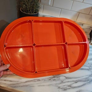 May include: A bright orange oval serving tray featuring six distinct compartments. The tray includes two large central square sections, two semi-circular side sections, and two smaller corner sections, all integrated into a single moulded plastic unit.