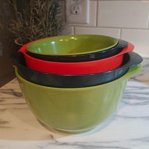 May include: A set of four nested mixing bowls in varying sizes. The bowls feature a smooth finish and side handles, colored in alternating shades of light green, black, and red, stacked vertically from the largest green bowl at the base.