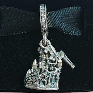 Puede incluir: Un charm de plata que presenta una estructura de castillo detallada adornada con pequeñas estrellas y una figura de hada sosteniendo una varita. El pasador está adornado con una hilera de piedras claras. El diseño incorpora pequeños acentos azules.