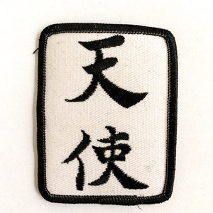 Puede incluir: Un parche de tela blanca con caracteres chinos bordados en negro que se leen "天" y "像". El parche tiene un borde negro.