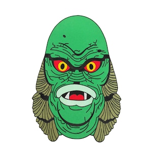 May include: An embroidered patch featuring a green creature face with yellow eyes, red pupils, and a wide mouth showing white teeth. The creature has textured skin lines and tan gill-like structures on both sides of its head, outlined in black.