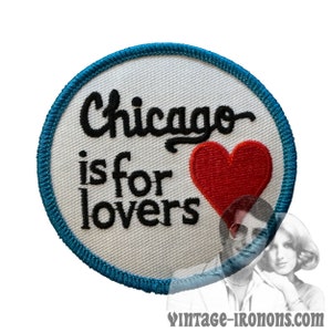 Puede incluir: Un parche bordado blanco con un borde azul. El parche presenta el texto "Chicago is for lovers" en fuente cursiva negra y un corazón rojo.