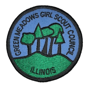 Op de afbeelding: Geborduurde patch van de Green Meadows Girl Scout Council Illinois met een blauwe achtergrond, groene bomen en zwarte tekst.