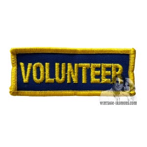 Può includere: Una toppa ricamata blu e oro con la parola "VOLUNTEER" in lettere gialle.