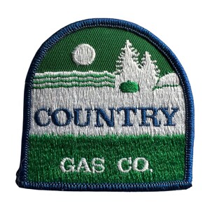 Op de afbeelding: Een halfronde geborduurde patch met een groen landschap met witte dennenbomen en een witte zon. De tekst "COUNTRY" is in het blauw in het midden geborduurd en "GAS CO." in het wit onderaan, alles omrand met een blauwe boord.