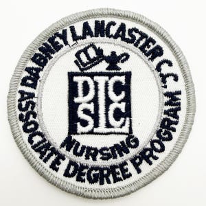 May include: A circular embroidered patch with a white background and a gray stitched border. The text reads Dabney Lancaster Community College Associate Degree Program Nursing. A central black square features the letters DLCC and a lamp icon.
