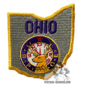 Może przedstawiać: Naszywka haftowana w kolorze szarym i żółtym w kształcie stanu Ohio. Naszywka zawiera napis "OHIO" w kolorze fioletowym i fioletowo-złoty okrąg z jeleniem i tarczą zegara. Tarcza zegara ma cyfry rzymskie i napisy "Buckeye" i "State" wokół krawędzi.