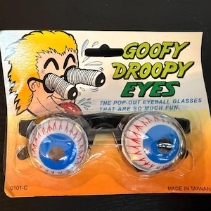 May include: Goofy Droopy Eyes pop-out eyeball glasses. The glasses have large, cartoonish eyeballs with blue irises and red veins. The packaging includes a cartoon illustration and the text "GOOFY DROOPY EYES".