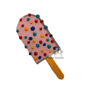 Könnte beinhalten: Ein gestickter Aufnäher in Form eines Eis am Stiel mit einer hellrosa Oberseite, die mit verstreuten, bunten Punkten in Blau, Gelb und Rot versehen ist. Das Eis hat einen strukturierten gelben Stiel und einen grauen Rand.