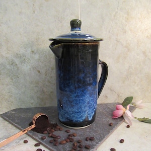 May include: A ceramic French press with a dark blue and black glaze. The coffee press is on a slate tile with coffee beans and a measuring scoop. A pink flower is in the background.