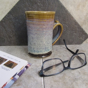 May include: A ceramic mug with a brown, purple, and blue glaze. The mug has a handle and a wide base. The mug is sitting on a table next to a pair of glasses and a book.