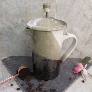 May include: A ceramic French press coffee maker with a brown, white, and black glaze. The coffee maker has a lid with a knob on top and a handle on the side. Coffee beans are scattered around the coffee maker.