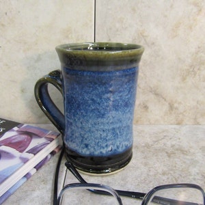 May include: A handmade ceramic mug with a blue and black glaze. The mug has a dark green rim and a curved handle. A pair of eyeglasses and a book are in the foreground.