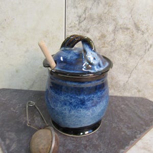 May include: A blue and white ceramic honey pot with a wooden dipper and a metal strainer. The pot has a brown handle and a black rim.