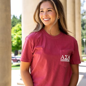 May include: A red short-sleeved t-shirt with a pocket on the left side. The pocket has white text that reads "AΣA ALPHA SIGMA ALPHA".