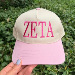 May include: A baseball cap with a cream-colored crown and a light pink brim. The front of the crown features the word ZETA embroidered in bright pink capital letters. The cap has a structured five-panel design with a matching pink button on top.