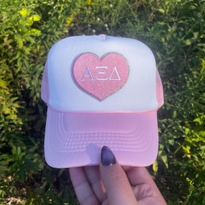 May include: A pink and white trucker hat with a pink heart patch that says "ΑΞΔ" in white letters.