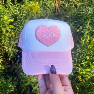 May include: A pink and white trucker hat with a pink heart embroidered with the letters "AΦ" in white.
