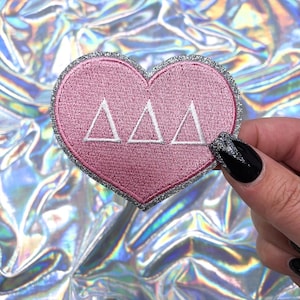 May include: Pink heart-shaped patch with a silver glitter border and white embroidered letters spelling "ΔΔΔ".