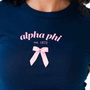 May include: A navy blue t-shirt with the text "alpha phi est. 1872" and a pink bow.
