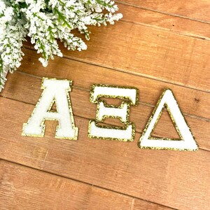 May include: Three white felt letters with gold glitter trim spelling out "AEA". The letters are on a woodgrain background.