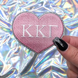 May include: Pink heart-shaped patch with silver glitter trim and white embroidered letters spelling "KKT".