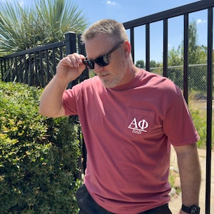 May include: A red-pink short-sleeved t-shirt with a pocket on the chest. The pocket features white Greek letters and the word "DAD" underneath. The person in the image is wearing sunglasses.