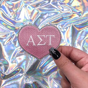 May include: A pink heart-shaped patch with the Greek letters ΑΣΤ embroidered in white. The patch has a silver glitter border. The background is a holographic, iridescent material.