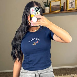 May include: A navy blue t-shirt with a white embroidered design featuring the text "ALPHA PHI" and a small teddy bear holding an American flag. The text "18 72" is also embroidered below the bear.