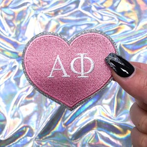 May include: Pink heart-shaped patch with silver glitter trim and white embroidered letters "AΦ" in the center.