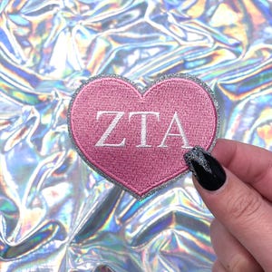 May include: A pink heart-shaped patch with silver glitter trim and the letters "ZTA" in white.