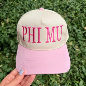 May include: A cream-colored baseball cap featuring the text PHI MU embroidered in bright pink thread across the front panel. The cap has a solid light pink brim with visible stitching and a matching pink button at the top center of the crown.