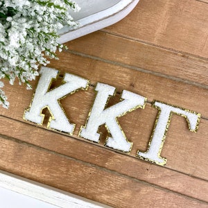 May include: White felt letters with gold glitter trim spelling out "KKT" on a wood background.