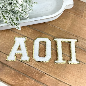 May include: Three white felt letters with gold glitter trim spelling out "AOII" on a wooden surface. The letters are in a collegiate style, with a textured appearance. A white decorative tray with greenery is in the background.