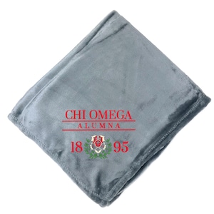 May include: A folded, gray fleece blanket with the words "CHI OMEGA ALUMNA" in red, along with the numbers "1895" and a floral emblem. The blanket is a soft, plush material.