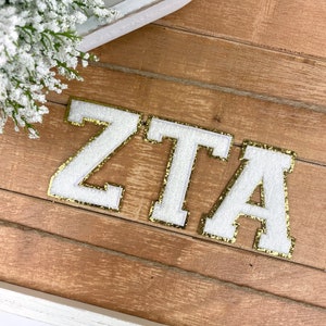 May include: White felt letters spelling "ZTA" with gold glitter trim on a wooden background.