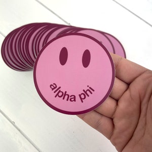 May include: Pink and burgundy smiley face sticker with the text "alpha phi" in burgundy.