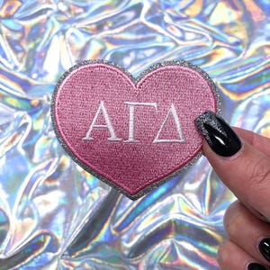 May include: Pink heart-shaped patch with silver glitter trim and white embroidered letters spelling "ΑΓΔ".