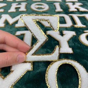 May include: White felt letter "Σ" with a gold glitter outline. The letter is on a green background with other white felt letters.