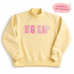 May include: A pale yellow mock neck sweatshirt featuring the text SIG KAP in pink letters with a mix of solid, gingham, and floral patterns. A pink speech bubble in the upper right corner contains the text available in 20+ Sororities!
