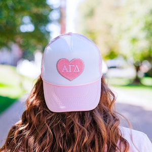 May include: A pink and white trucker hat with a pink heart featuring the letters "AGD" in white.