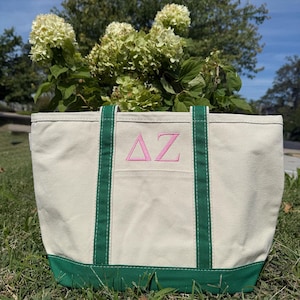 May include: A canvas tote bag with a cream-colored body, green trim, and handles. The front features a pink embroidered "ΔZ" design. The bag is sitting on grass with a backdrop of green foliage and a blue sky.